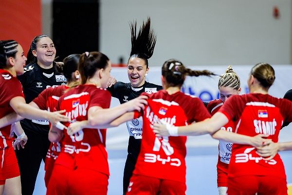 A group of athletes in red uniforms celebrate together on a court, with one player joyfully raising her hair as teammates embrace and cheer.